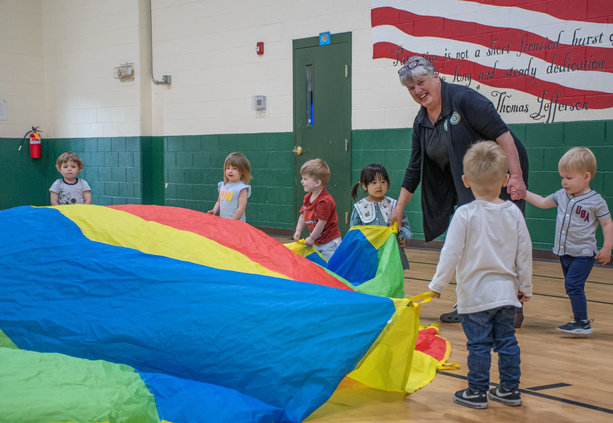 A teacher and young students smiling while holding and playing with a large colorful parachute game indoors in a gymnasium.