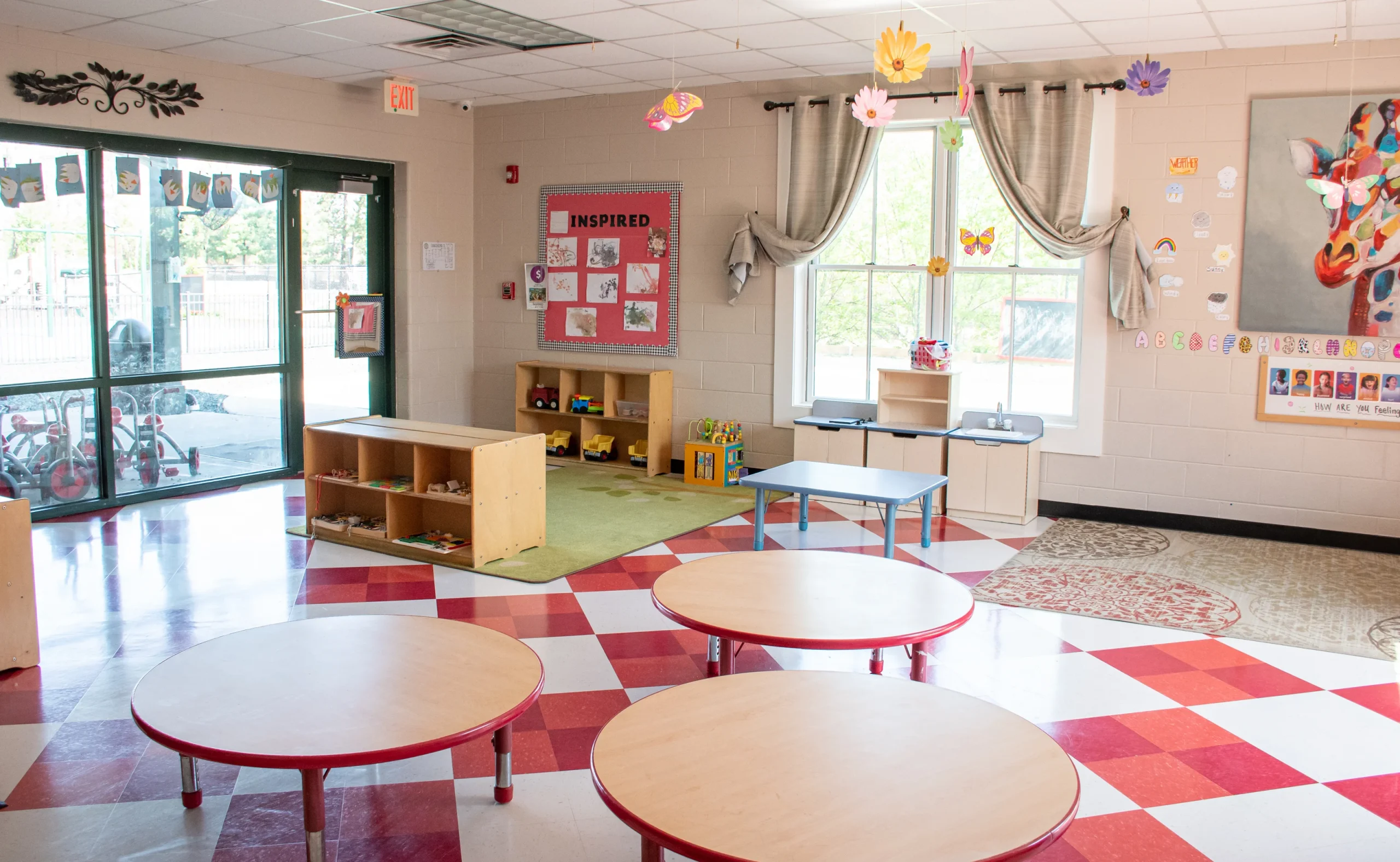 An interior shot of a brightly lit daycare activity space featuring circular wooden tables, learning materials on low shelves, and large windows.