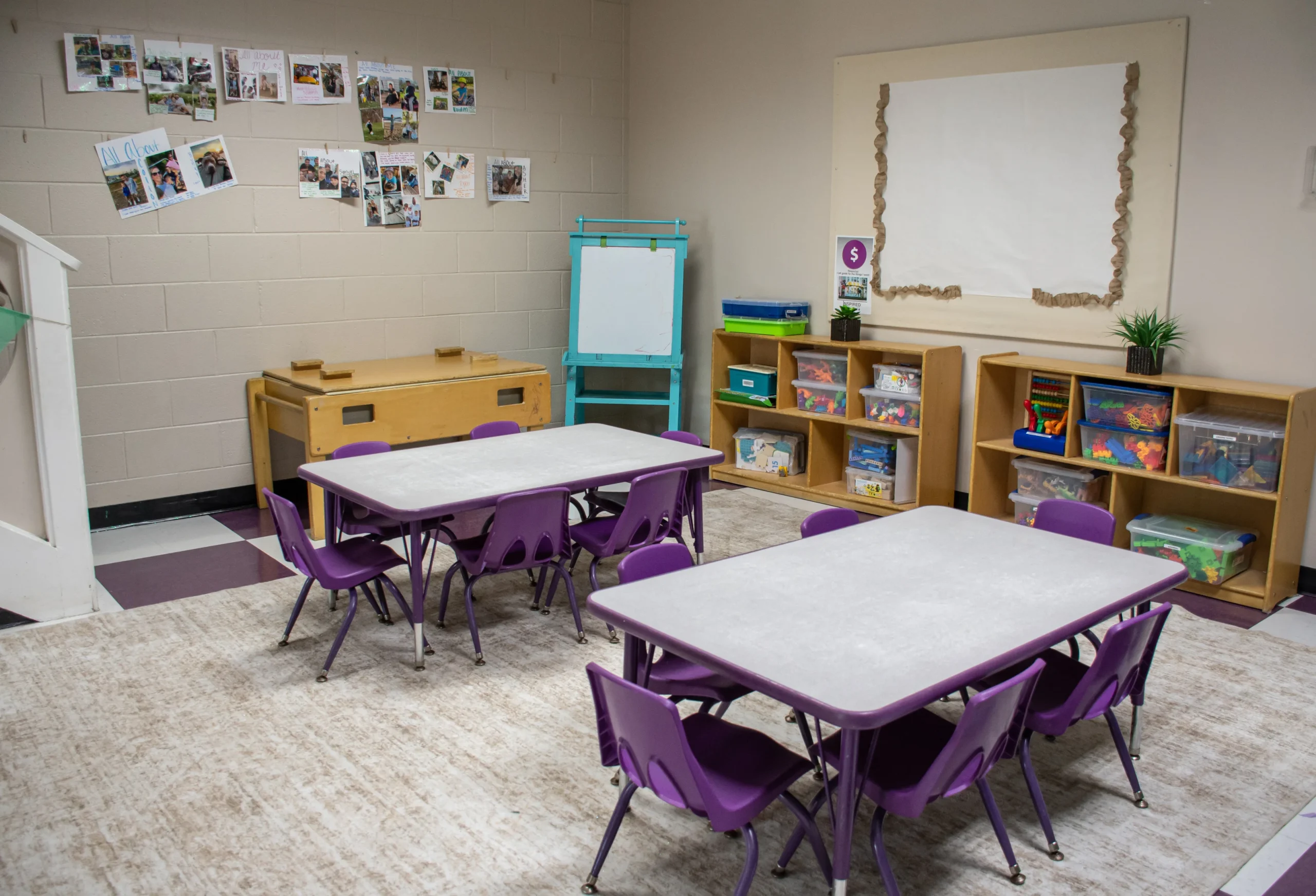 An interior view of an empty preschool classroom featuring small purple chairs, activity tables, shelving units filled with toys, and educational wall displays.